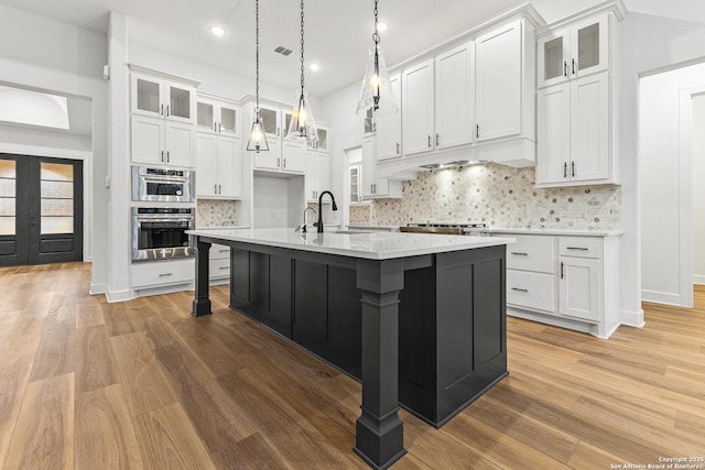 kitchen with glass insert cabinets, a kitchen island with sink, white cabinetry, and light countertops