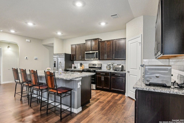 kitchen with dark wood-type flooring, a breakfast bar, appliances with stainless steel finishes, light stone countertops, and a kitchen island with sink