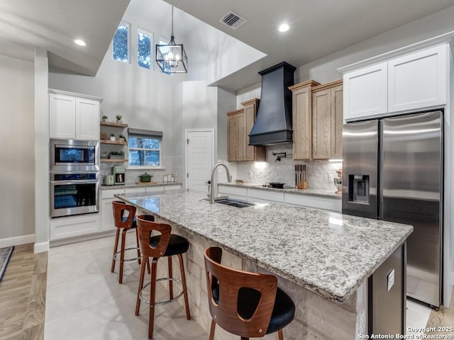 kitchen with sink, custom range hood, stainless steel appliances, a kitchen island with sink, and white cabinets