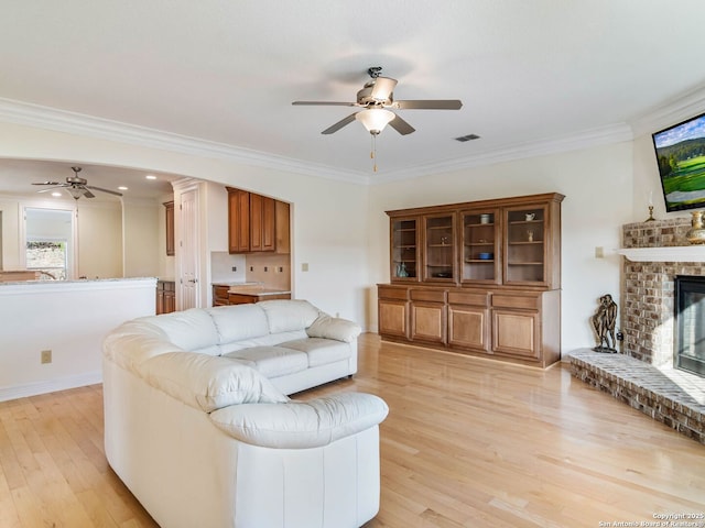 living room featuring ceiling fan, ornamental molding, light hardwood / wood-style floors, and a brick fireplace