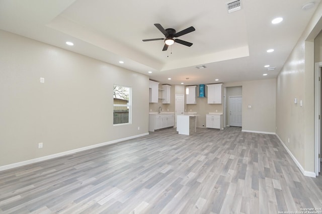 unfurnished living room with ceiling fan, a tray ceiling, sink, and light wood-type flooring