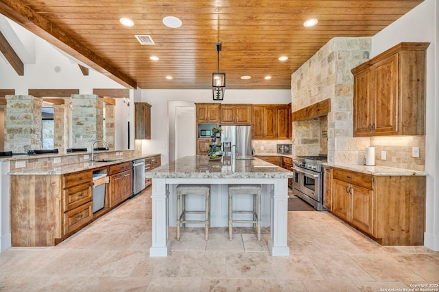 kitchen with an island with sink, appliances with stainless steel finishes, light stone counters, and decorative light fixtures