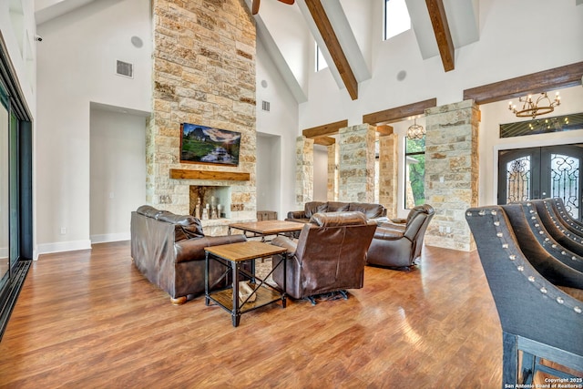 living room with beam ceiling, a notable chandelier, wood-type flooring, a stone fireplace, and french doors