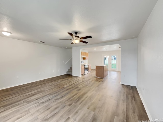 unfurnished living room featuring arched walkways, stairway, light wood-style flooring, ceiling fan, and baseboards