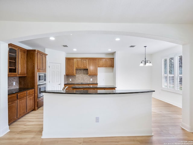 kitchen featuring light wood-type flooring, a kitchen island with sink, stainless steel appliances, hanging light fixtures, and dark stone countertops