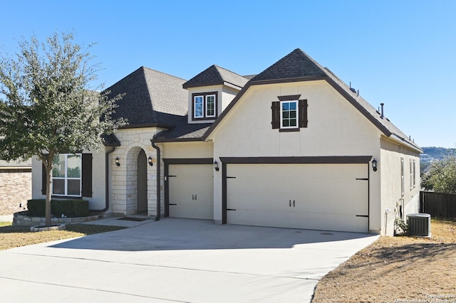 french country inspired facade featuring roof with shingles, stucco siding, central air condition unit, stone siding, and driveway