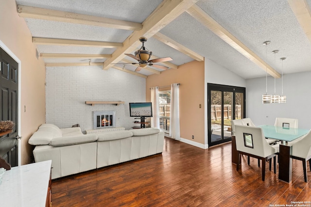 living area featuring ceiling fan with notable chandelier, lofted ceiling with beams, a textured ceiling, dark wood-style floors, and a brick fireplace