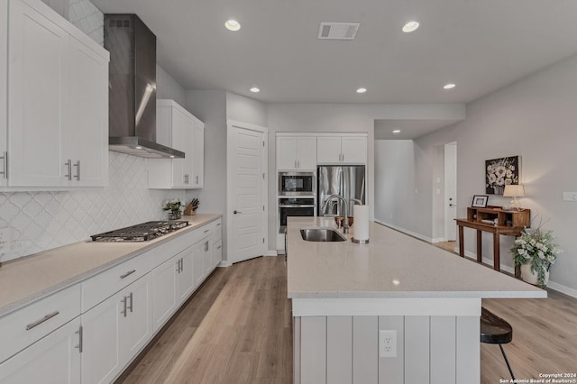 kitchen with white cabinetry, a center island with sink, sink, appliances with stainless steel finishes, and wall chimney range hood