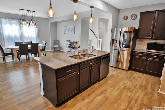 kitchen with dark brown cabinets, stainless steel appliances, pendant lighting, light wood-type flooring, and a kitchen island with sink