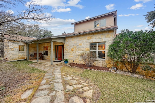 rear view of house with stone siding, stucco siding, a lawn, and a patio