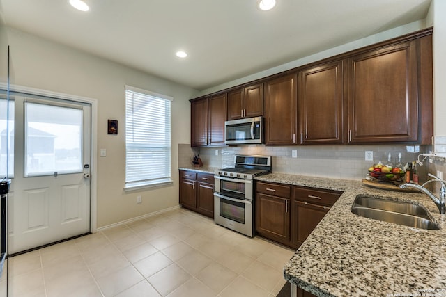 kitchen featuring dark brown cabinetry, stainless steel appliances, a sink, light stone countertops, and tasteful backsplash