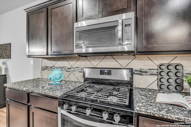 kitchen with gas range, stainless steel microwave, dark stone countertops, dark brown cabinets, and backsplash