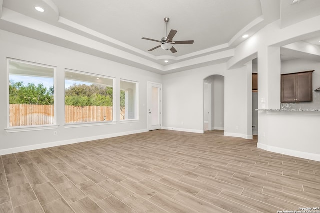 unfurnished living room featuring arched walkways, a raised ceiling, a ceiling fan, light wood-type flooring, and baseboards