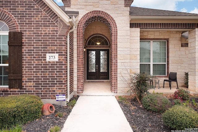 view of exterior entry featuring french doors, a shingled roof, and brick siding