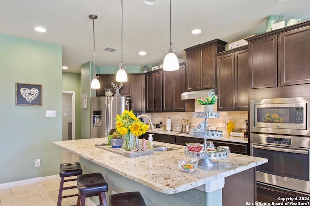 kitchen featuring light stone counters, hanging light fixtures, appliances with stainless steel finishes, dark brown cabinetry, and an island with sink