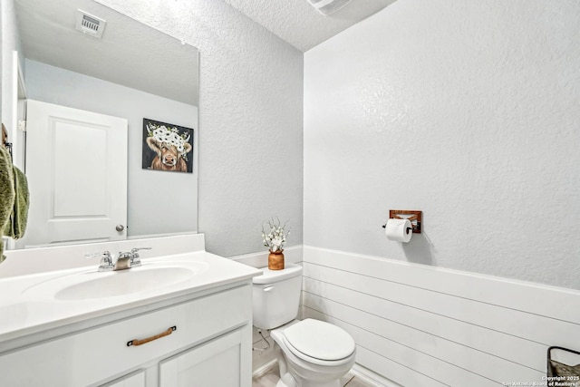 bathroom featuring a textured wall, toilet, vanity, visible vents, and wainscoting