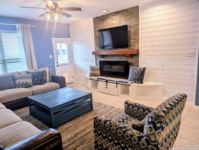 living area featuring a ceiling fan, wood walls, a stone fireplace, and light tile patterned floors