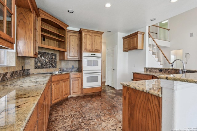 kitchen with light stone counters, brown cabinets, white double oven, backsplash, and glass insert cabinets