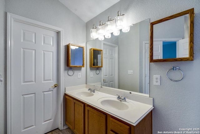 full bath featuring a textured wall, vaulted ceiling, a sink, and double vanity