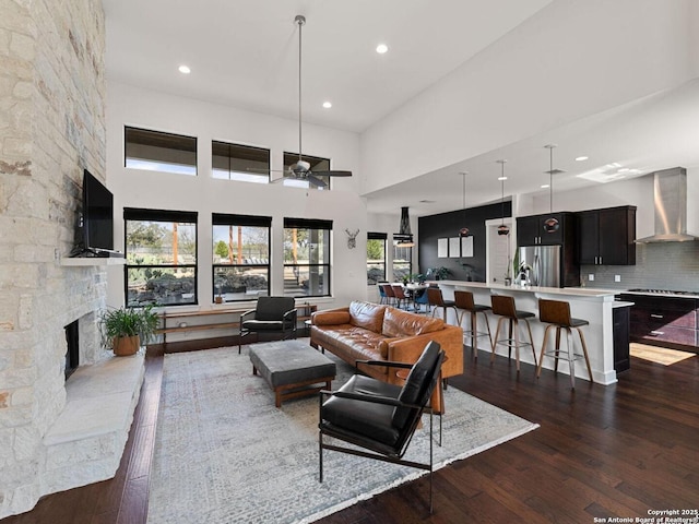 living room featuring recessed lighting, a towering ceiling, dark wood-type flooring, a ceiling fan, and a stone fireplace