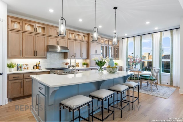 kitchen featuring decorative light fixtures, light countertops, glass insert cabinets, an island with sink, and under cabinet range hood