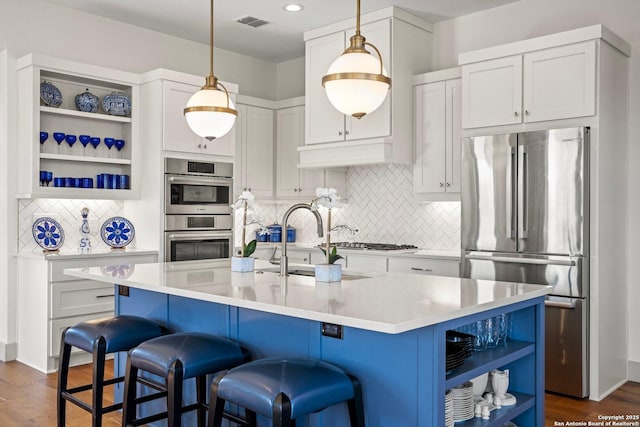 kitchen featuring decorative light fixtures, a kitchen island with sink, stainless steel appliances, open shelves, and a sink