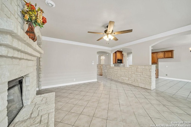 unfurnished living room featuring ceiling fan, light tile patterned floors, a stone fireplace, baseboards, and crown molding