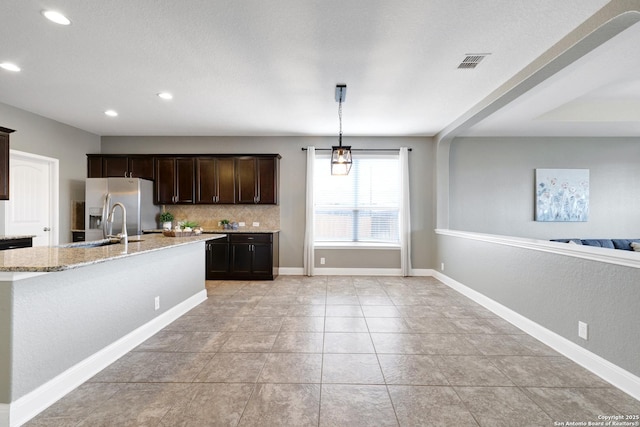 kitchen with light stone counters, pendant lighting, a center island with sink, stainless steel refrigerator with ice dispenser, and open floor plan