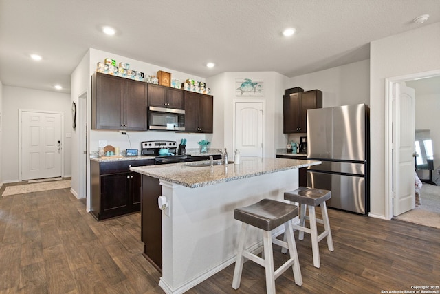 kitchen featuring an island with sink, dark wood-style floors, stainless steel appliances, dark brown cabinets, and a sink