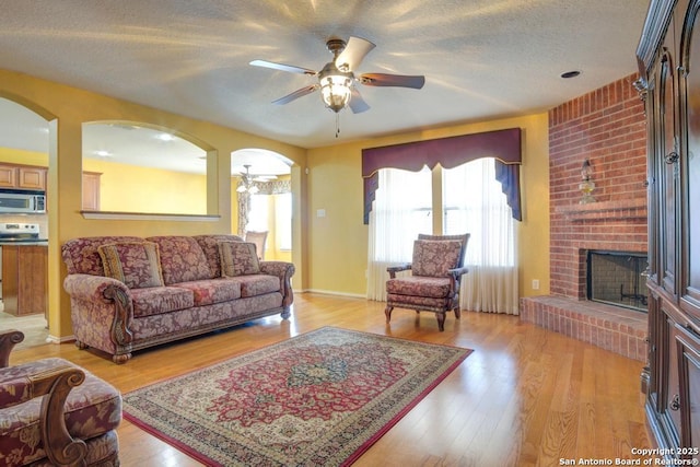 living room with a brick fireplace, arched walkways, a ceiling fan, a textured ceiling, and light wood-style floors