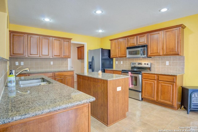 kitchen with a center island, stainless steel appliances, brown cabinetry, a sink, and light stone countertops