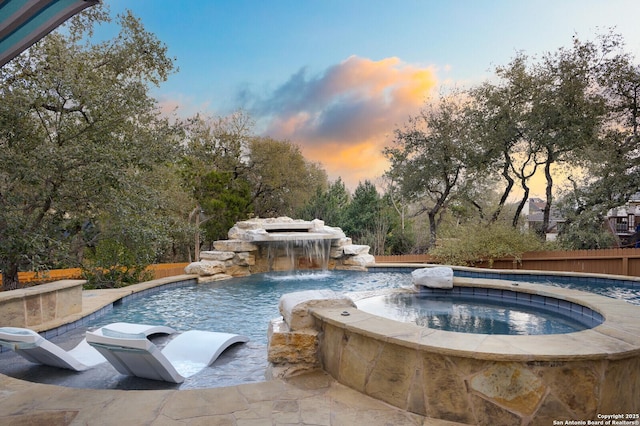 pool at dusk with an outdoor pool, fence, and an in ground hot tub