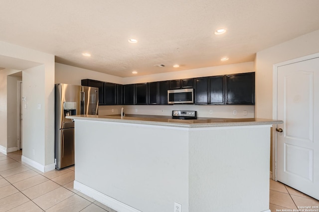 kitchen with light tile patterned floors, dark cabinets, stainless steel appliances, light countertops, and an island with sink