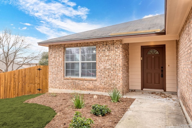 property entrance featuring brick siding, fence, and roof with shingles