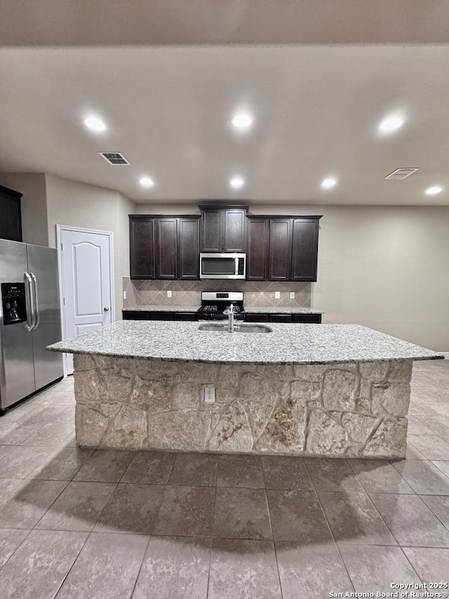 kitchen featuring appliances with stainless steel finishes, a kitchen island with sink, dark brown cabinetry, and light stone counters