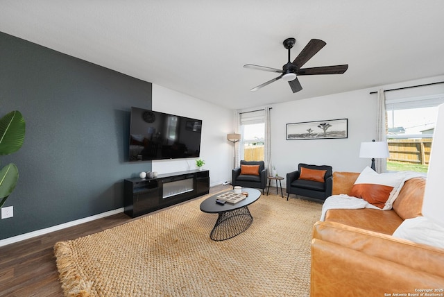 living area featuring a glass covered fireplace, dark wood finished floors, baseboards, and ceiling fan