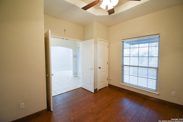 unfurnished bedroom with arched walkways, a raised ceiling, dark wood finished floors, and baseboards