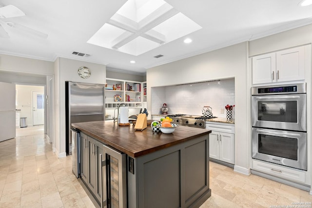 kitchen with a center island with sink, butcher block counters, stainless steel appliances, crown molding, and white cabinetry