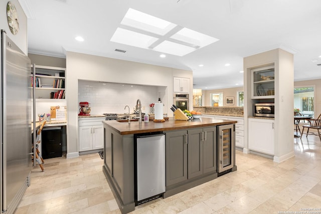 kitchen with a center island with sink, stainless steel appliances, white cabinetry, wood counters, and beverage cooler