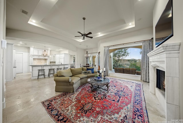 living room with ceiling fan with notable chandelier, a tray ceiling, visible vents, and a premium fireplace