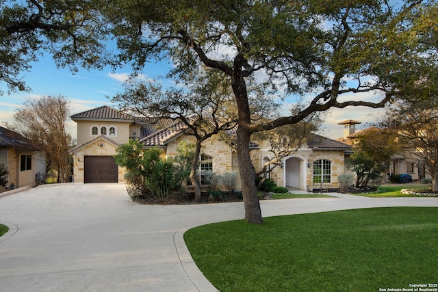 mediterranean / spanish house with driveway, stone siding, a tiled roof, an attached garage, and a front lawn