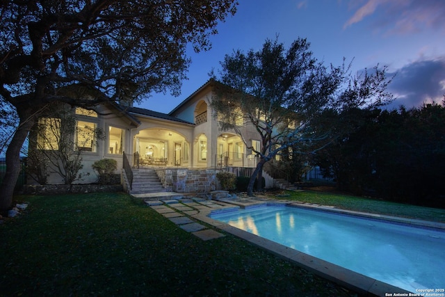 back of house at dusk featuring a fenced in pool, a yard, stairway, and stucco siding