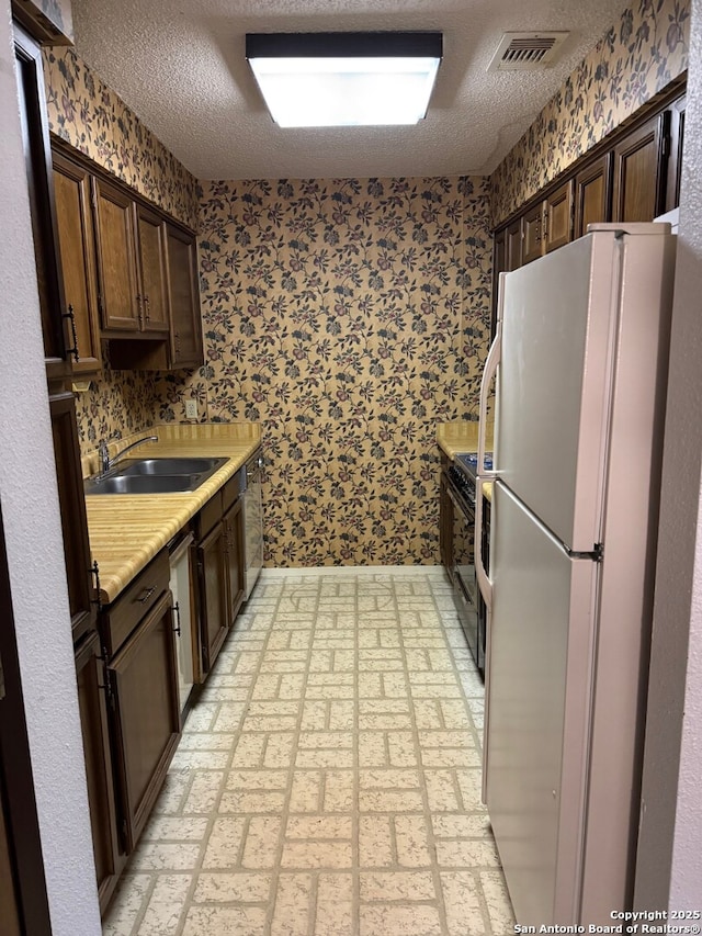 kitchen featuring dark brown cabinetry, a sink, light countertops, freestanding refrigerator, and wallpapered walls