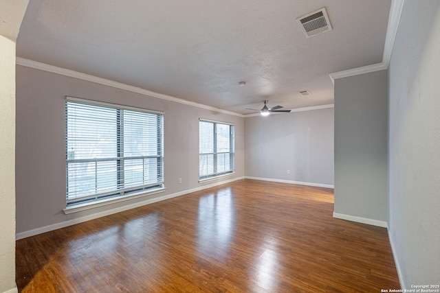 empty room with wood finished floors, visible vents, a ceiling fan, crown molding, and baseboards
