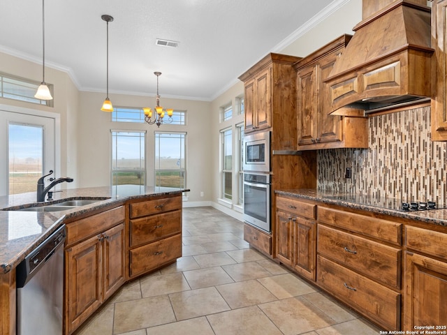 kitchen with dark stone counters, custom range hood, appliances with stainless steel finishes, decorative light fixtures, and a sink