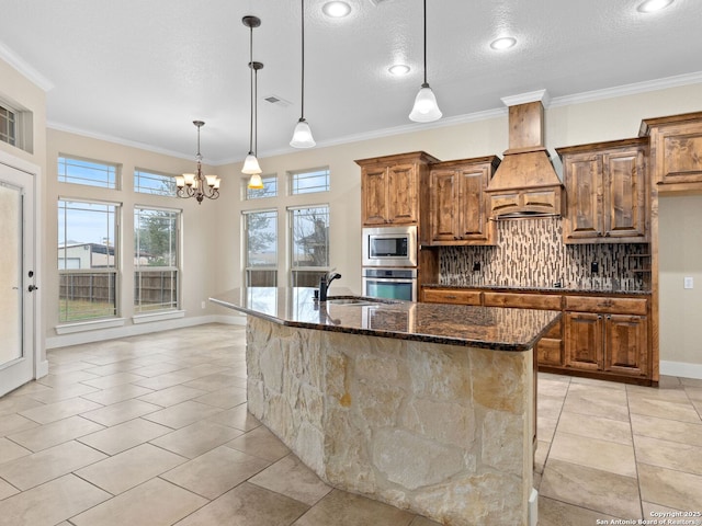 kitchen with a center island with sink, dark stone counters, decorative light fixtures, stainless steel appliances, and premium range hood