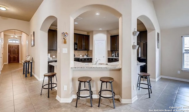 kitchen with dark brown cabinetry, tasteful backsplash, arched walkways, a kitchen breakfast bar, and light stone countertops