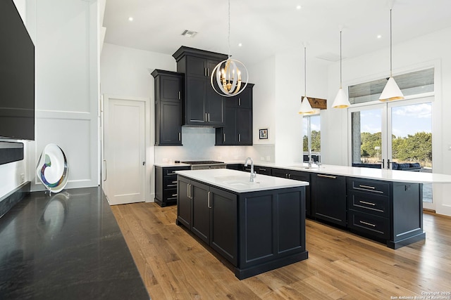 kitchen featuring a center island with sink, a chandelier, a sink, wood finished floors, and dark cabinets