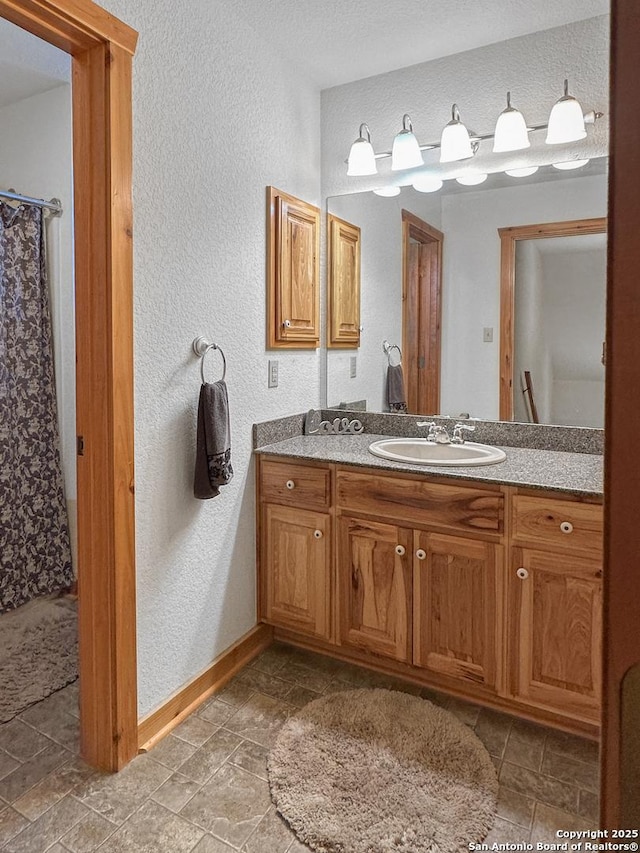 full bathroom with stone finish floor, a textured wall, vanity, and baseboards