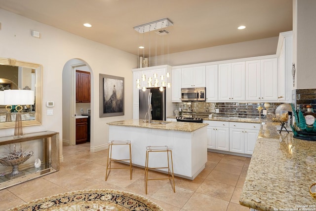 kitchen with an island with sink, appliances with stainless steel finishes, hanging light fixtures, light stone countertops, and white cabinetry